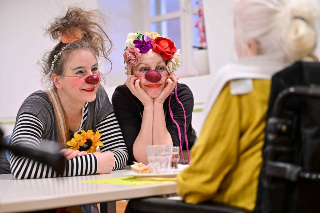 Deux femmes habillées en clowns avec des nez rouges et des accessoires colorés sourient et s'appuient sur une table, s'engageant chaleureusement avec une personne âgée en fauteuil roulant, vue de dos.