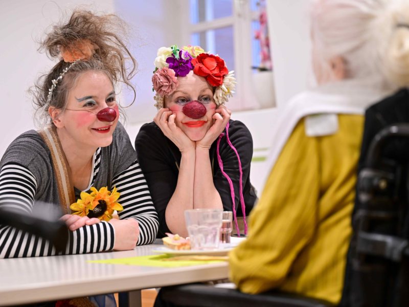 Deux femmes habillées en clowns avec des nez rouges et des accessoires colorés sourient et s'appuient sur une table, s'engageant chaleureusement avec une personne âgée en fauteuil roulant, vue de dos.