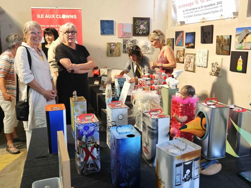 Un groupe de personnes se tient autour d'une table où sont exposées des boîtes de conserve colorées et peintes, ainsi que des œuvres d'art mixtes, dans un marché d'art intérieur. Diverses peintures et petites œuvres d'art décorent le mur derrière la table.