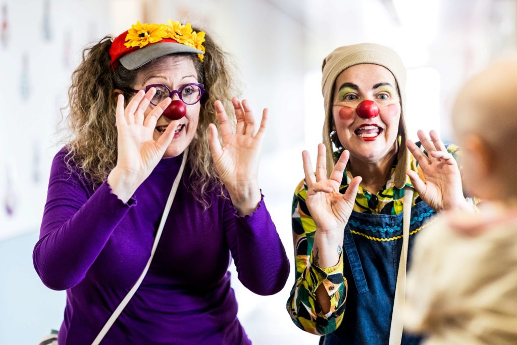 Deux femmes habillées en clowns, avec des nez rouges, des maquillages colorés et des expressions enjouées, amusent un enfant. L'une porte un bandeau de fleurs et des lunettes, l'autre un chapeau. Toutes deux lèvent les mains, sourient et font des gestes ridicules.