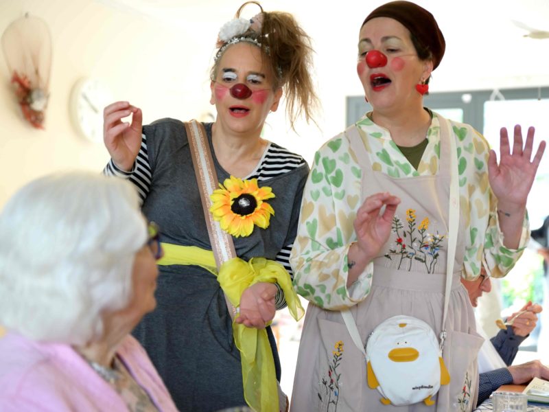 Deux femmes habillées en clowns avec des nez rouges et des costumes colorés interagissent joyeusement avec une femme âgée assise, créant une atmosphère ludique et joyeuse dans un intérieur lumineux.