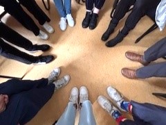 Un groupe de personnes debout en cercle, photographiées de haut, ne montrant que leurs jambes et divers types de chaussures sur un sol beige.