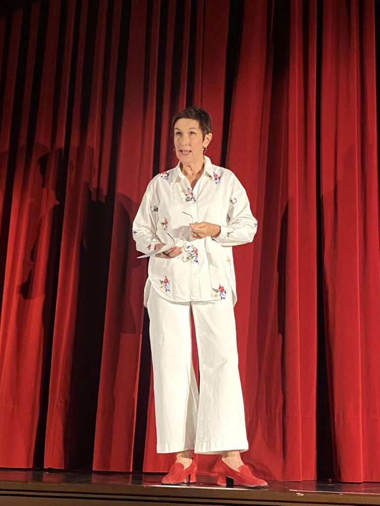 Une personne aux cheveux courts se tient sur scène devant des rideaux rouges, vêtue d'une chemise blanche à manches longues avec des broderies colorées, d'un pantalon blanc et de chaussures rouges, semblant parler ou jouer.