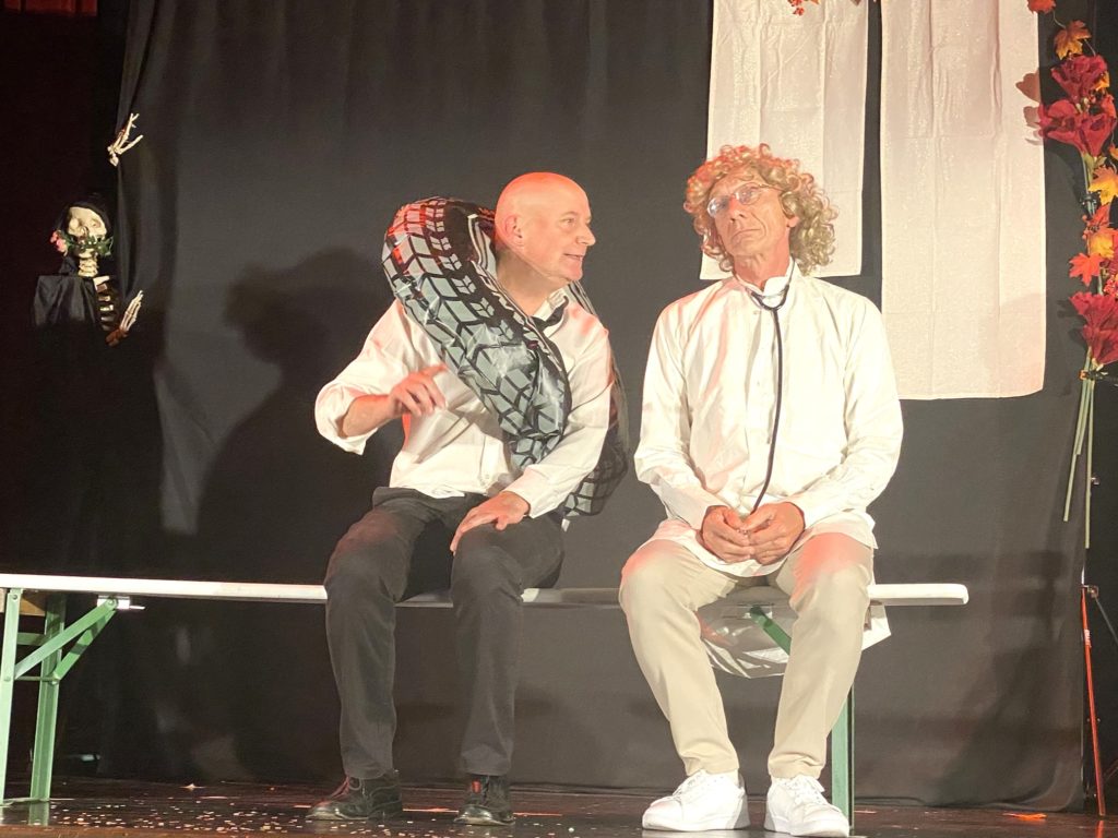 Deux hommes sont assis sur un banc sur scène ; l'un porte un pneu autour des épaules et fait des gestes tandis que l'autre, avec une perruque bouclée et des vêtements blancs, reste assis tranquillement. L'arrière-plan est décoré de fleurs et d'accessoires d'Halloween.