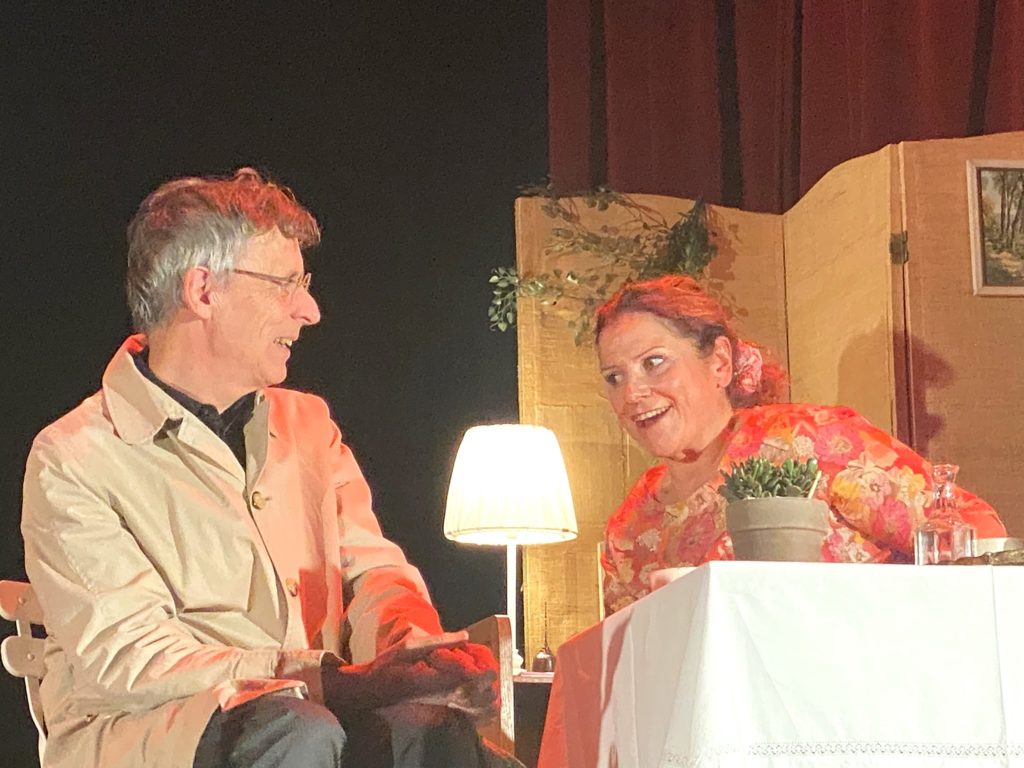 Un homme âgé vêtu d'un manteau beige et une femme portant un haut à fleurs sont assis à une petite table à l'intérieur, se souriant l'un à l'autre. Une lampe, une plante en pot et des bouteilles sont posées sur la table ; un écran et une photo encadrée se trouvent à l'arrière-plan.