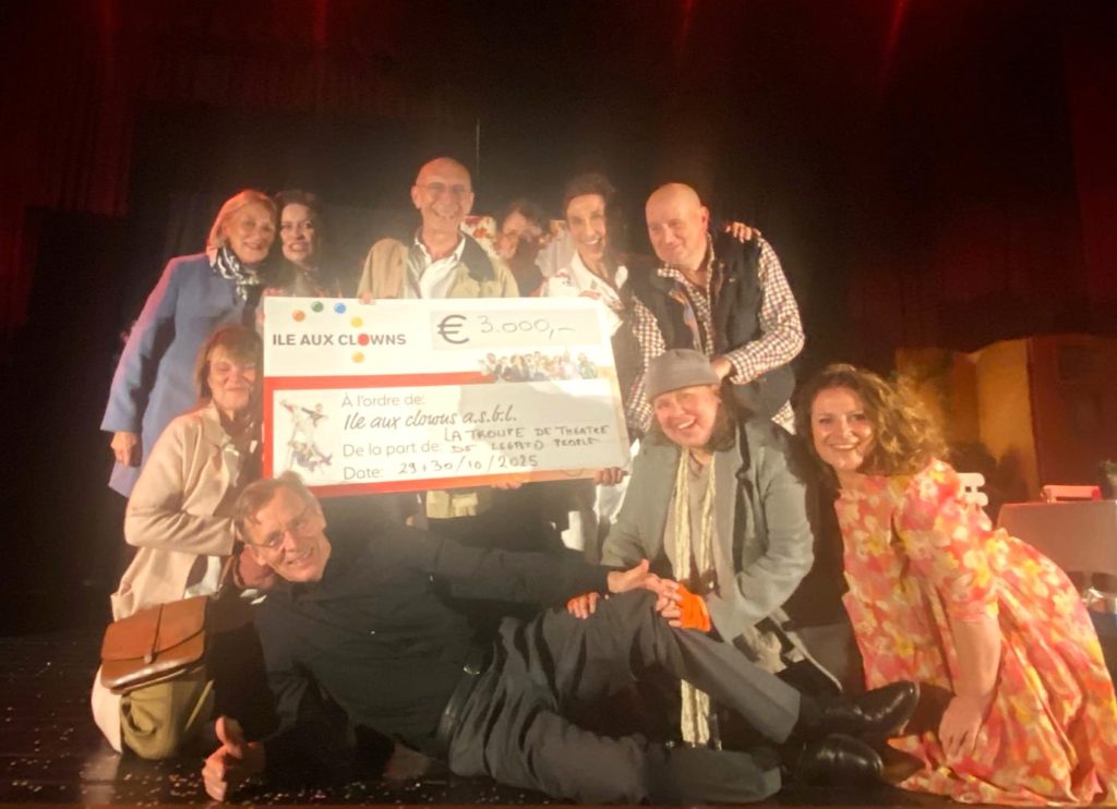 Un groupe de personnes souriant et posant sur scène, tenant un gros chèque de 3 000 euros à l'ordre de "Ile Aux Clowns", daté du 23/10/2023. L'arrière-plan est faiblement éclairé par des rideaux rouges.