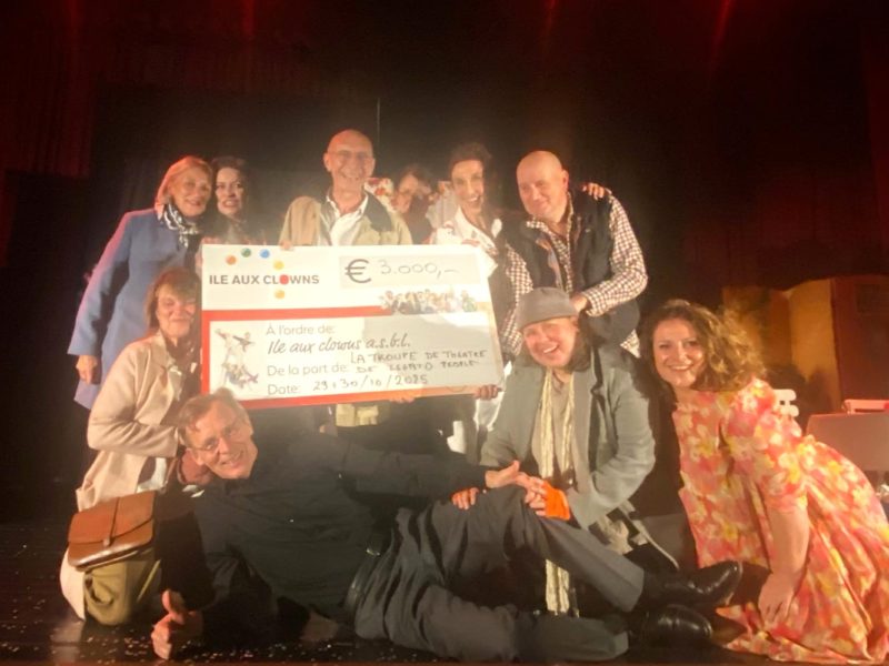 Un groupe de personnes souriant et posant sur scène, tenant un gros chèque de 3 000 euros à l'ordre de "Ile Aux Clowns", daté du 23/10/2023. L'arrière-plan est faiblement éclairé par des rideaux rouges.