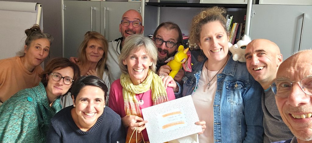 Un groupe de dix adultes souriants posent étroitement ensemble à l'intérieur, une personne tenant une carte et une autre une girafe jaune. Ils semblent heureux et joyeux dans un cadre décontracté.