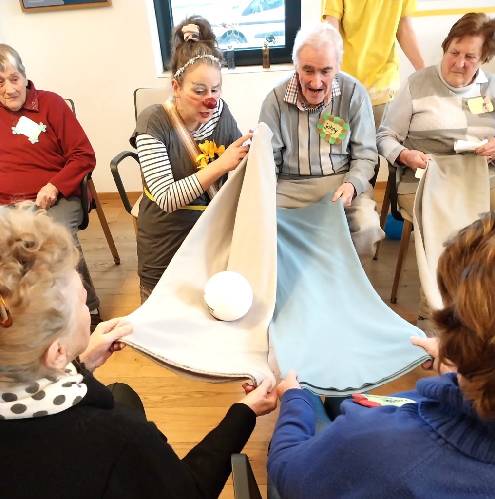 Un groupe de personnes âgées et un clown sont assis en cercle, souriant et utilisant de grands chiffons pour lancer une balle blanche entre eux, se livrant à une activité de groupe amusante en intérieur.