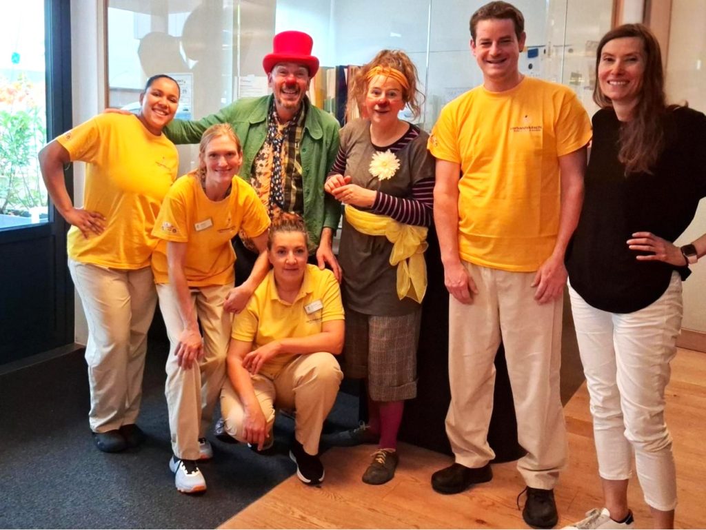 Six adultes se tiennent à l'intérieur et sourient à l'appareil photo. Quatre d'entre eux portent des chemises jaunes et des pantalons beiges, tandis que deux autres sont vêtus de costumes de clowns colorés et de chapeaux. Le décor semble lumineux et joyeux.