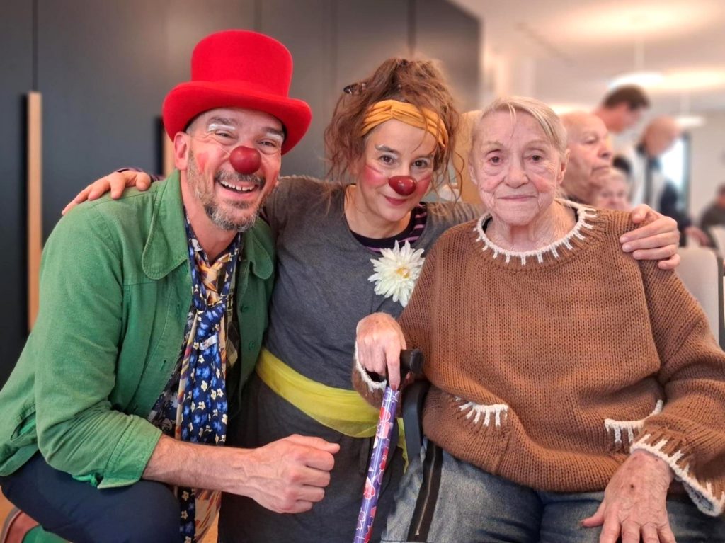 Trois personnes posent ensemble, deux habillées en clowns avec des nez rouges et des vêtements colorés, souriant, et une femme âgée avec une canne portant un pull marron, assise entre les deux, souriant également.
