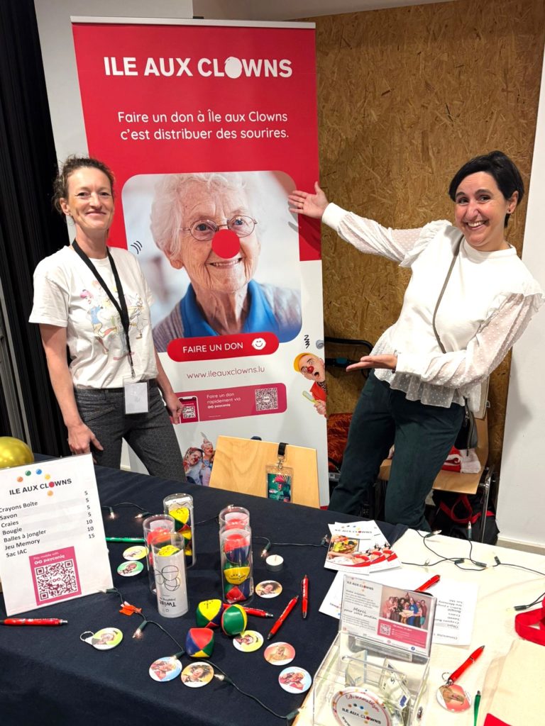 Deux femmes sourient devant une bannière de l'Île aux Clowns représentant une femme âgée avec un nez de clown. Une table à l'avant présente des articles colorés, des brochures et des informations sur les dons pour l'association caritative des clowns.