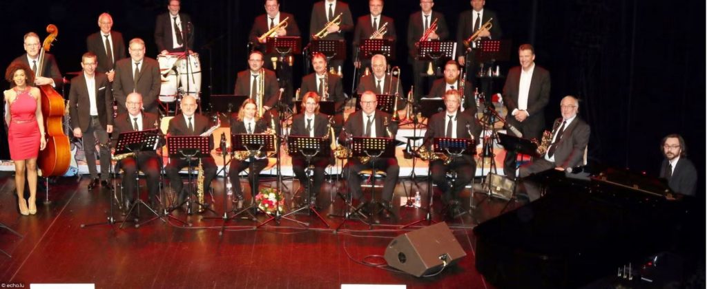 Un grand groupe de jazz en tenue de soirée pose sur scène avec divers instruments, dont des cuivres, des bois, une batterie, une guitare, une contrebasse et un piano. Une femme en robe rouge se tient à l'avant gauche. Des pupitres sont installés devant le groupe.