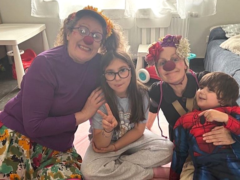 Deux adultes déguisés en clowns sont assis par terre avec deux enfants, l'un portant des lunettes et faisant un signe de paix, l'autre portant un costume de Spider-Man. Tout le monde sourit dans une pièce confortable et ensoleillée.