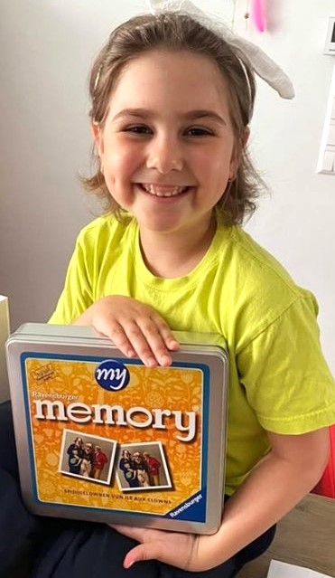 Un enfant souriant aux cheveux courts et au bandeau blanc tient une boîte en fer jaune étiquetée "ma mémoire" et est assis à l'intérieur. L'enfant porte un T-shirt jaune vif.