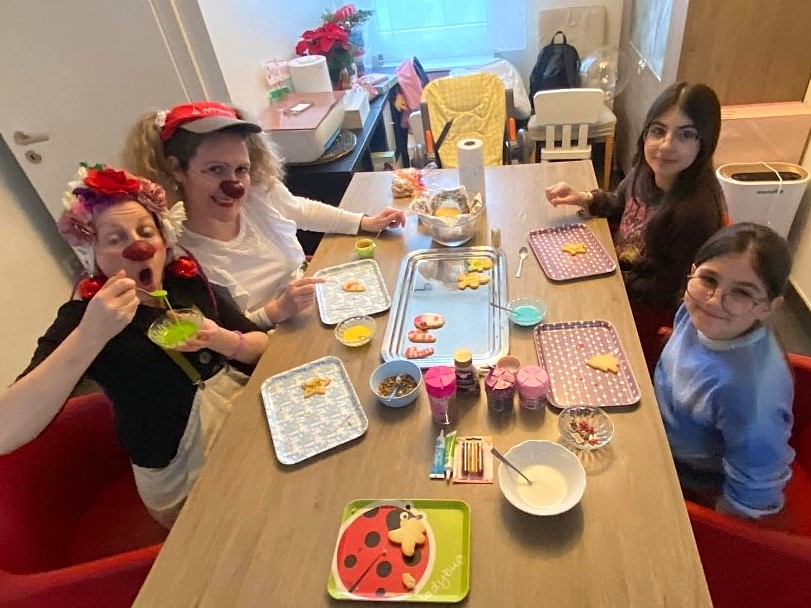 Quatre personnes, dont deux en costume de clown et maquillées, sont assises autour d'une table décorée avec du matériel de pâtisserie, des biscuits et du glaçage coloré, semblant décorer des biscuits ensemble dans une pièce confortable et lumineuse.