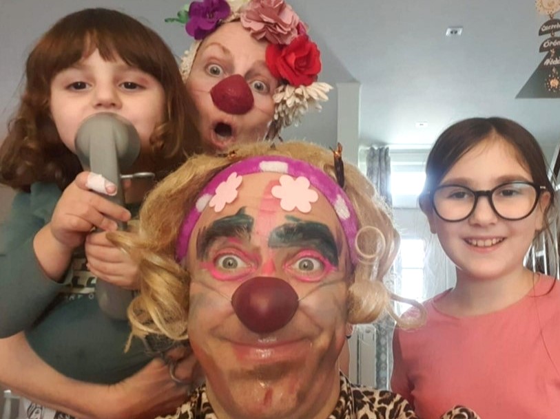 Trois personnes, dont deux habillées en clowns avec des visages peints, des nez rouges et des perruques, posent en jouant avec un enfant qui tient un sèche-cheveux jouet, et un autre enfant souriant portant des lunettes se tient à côté d'eux à l'intérieur.