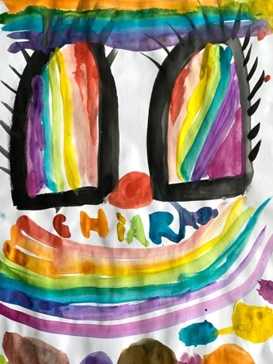 Peinture colorée et enfantine d'un visage aux yeux rayés d'arc-en-ciel, au nez rouge, au sourire arc-en-ciel et au nom Chiara écrit dans les couleurs de l'arc-en-ciel sous le nez et la bouche. Les yeux et les cils sont soulignés par des contours noirs.