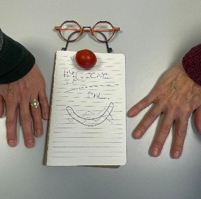 Deux mains reposent sur une table à côté d'un bloc-notes décoré de lunettes, d'un objet rond rouge en guise de nez et d'une bouche souriante dessinée, créant un arrangement en forme de visage sur le bloc-notes.