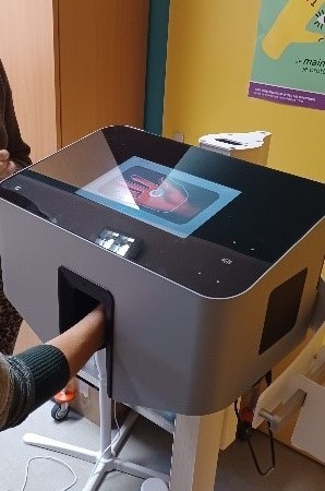 Une personne place son bras dans un dispositif médical gris doté d'un écran tactile numérique affichant l'image d'un organe interne, éventuellement à des fins de diagnostic ou de balayage.