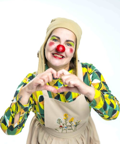 Une personne habillée en clown, portant un nez rouge, un maquillage coloré, une chemise à motifs verts et jaunes, un tablier beige avec des fleurs brodées et un chapeau beige, sourit et forme un cœur avec ses mains.