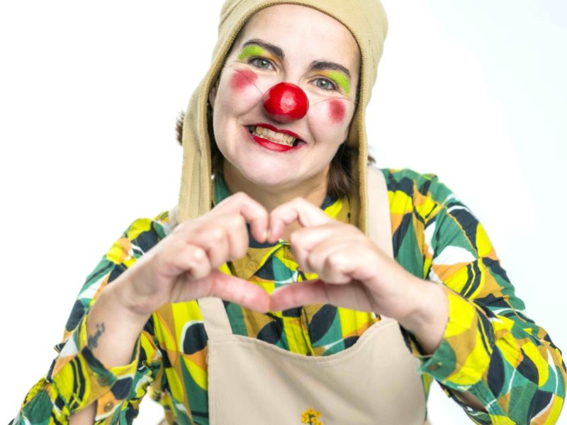 Une personne habillée en clown, portant un nez rouge, un maquillage coloré, une chemise à motifs verts et jaunes, un tablier beige avec des fleurs brodées et un chapeau beige, sourit et forme un cœur avec ses mains.