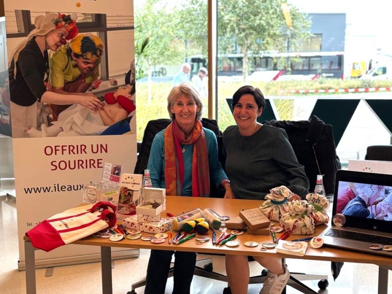 Deux femmes sont assises, souriantes, à une table où sont exposés des jouets et des objets colorés, avec une bannière "Ile Aux Clowns" derrière elles. La scène semble se dérouler à l'intérieur, près de grandes fenêtres, avec des arbres et des bâtiments visibles à l'extérieur.