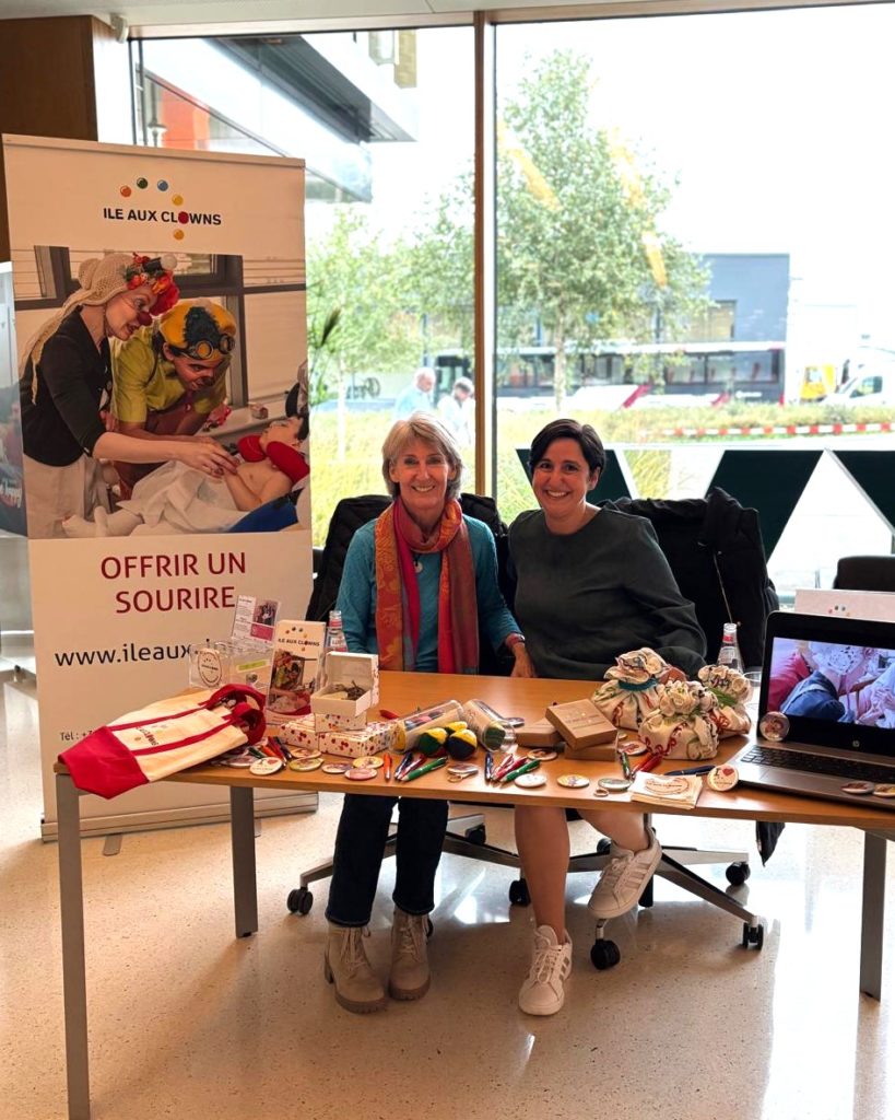 Deux femmes sont assises, souriantes, à une table où sont exposés des jouets et des objets colorés, avec une bannière "Ile Aux Clowns" derrière elles. La scène semble se dérouler à l'intérieur, près de grandes fenêtres, avec des arbres et des bâtiments visibles à l'extérieur.