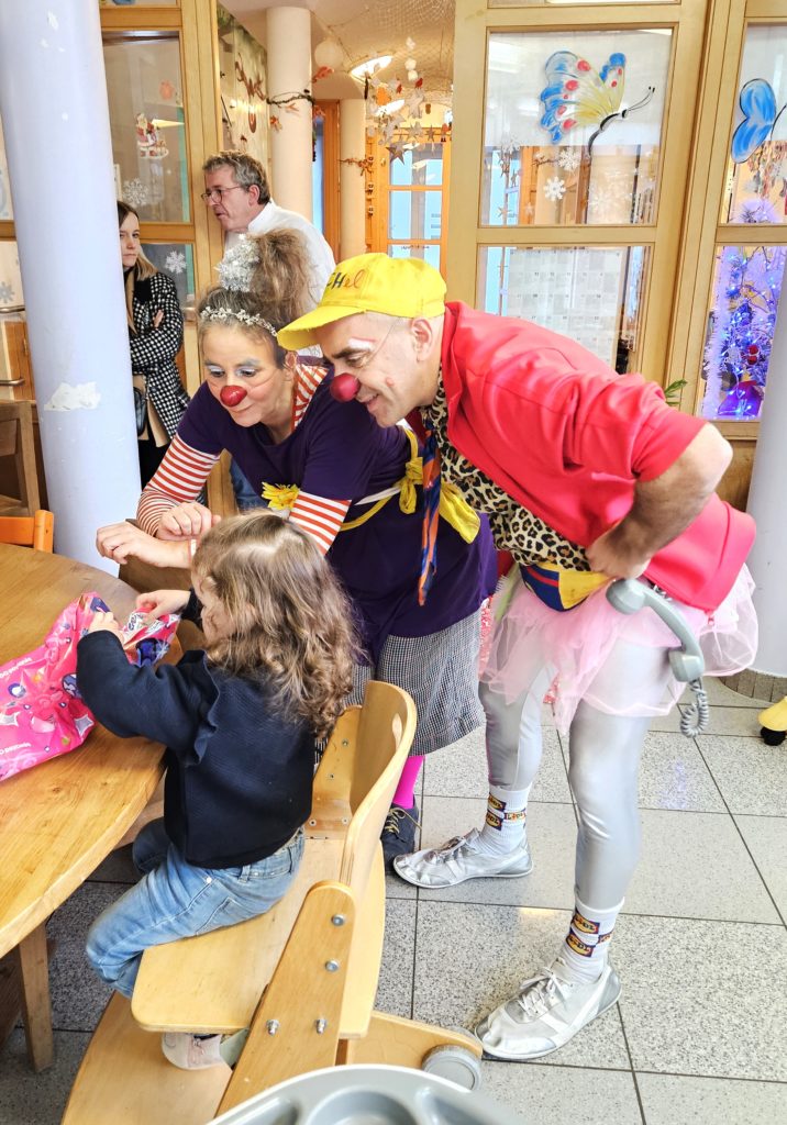 Deux clowns au nez rouge, aux tenues colorées et aux perruques amusent un jeune enfant assis à une table. D'autres adultes observent la scène en arrière-plan, dans un intérieur lumineux et décoré de papillons sur les murs.