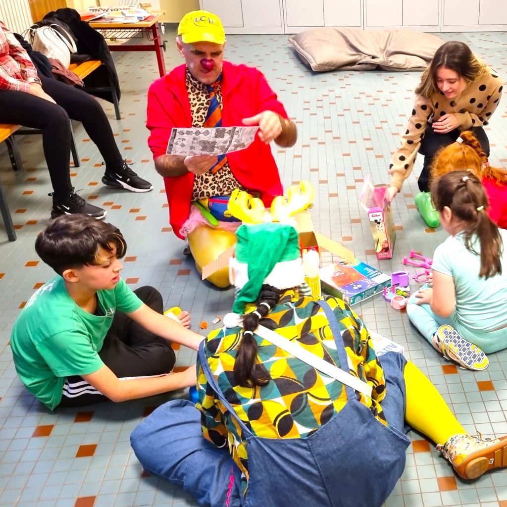 Un clown vêtu de vêtements colorés et coiffé d'un chapeau jaune est assis par terre avec des enfants, jouant avec des jouets et lisant un journal. D'autres enfants et un adulte se joignent à eux, profitant de l'activité dans une pièce bien éclairée.