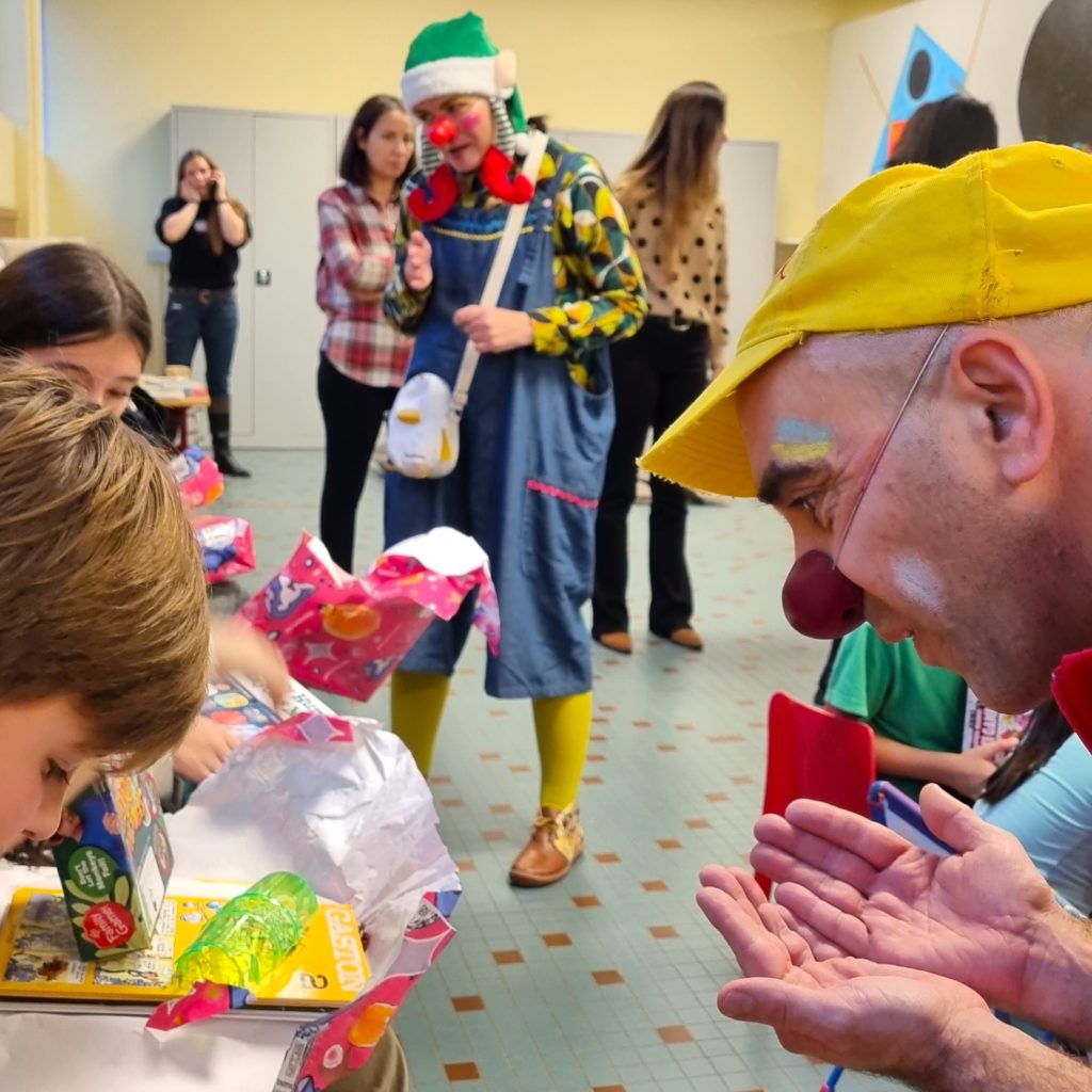 Un clown souriant coiffé d'une casquette jaune amuse des enfants qui ouvrent des cadeaux à une table, tandis qu'un autre clown et plusieurs adultes regardent à l'arrière-plan, dans une pièce très éclairée.