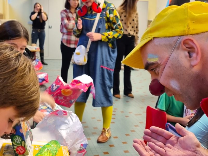 Un clown souriant coiffé d'une casquette jaune amuse des enfants qui ouvrent des cadeaux à une table, tandis qu'un autre clown et plusieurs adultes regardent à l'arrière-plan, dans une pièce très éclairée.