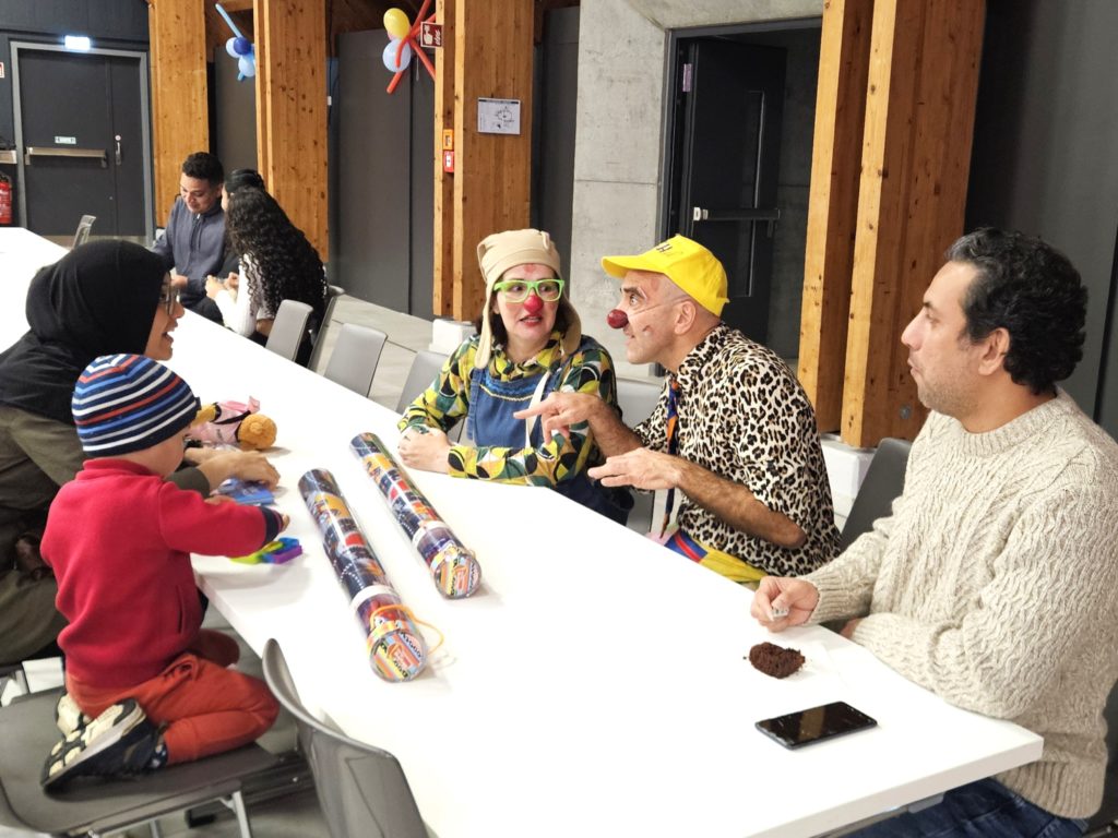Un groupe de personnes, dont deux clowns, sont assis autour d'une table blanche, discutant et jouant avec du matériel de bricolage, tandis qu'un enfant coiffé d'un chapeau coloré est assis à proximité ; la scène est animée et conviviale.