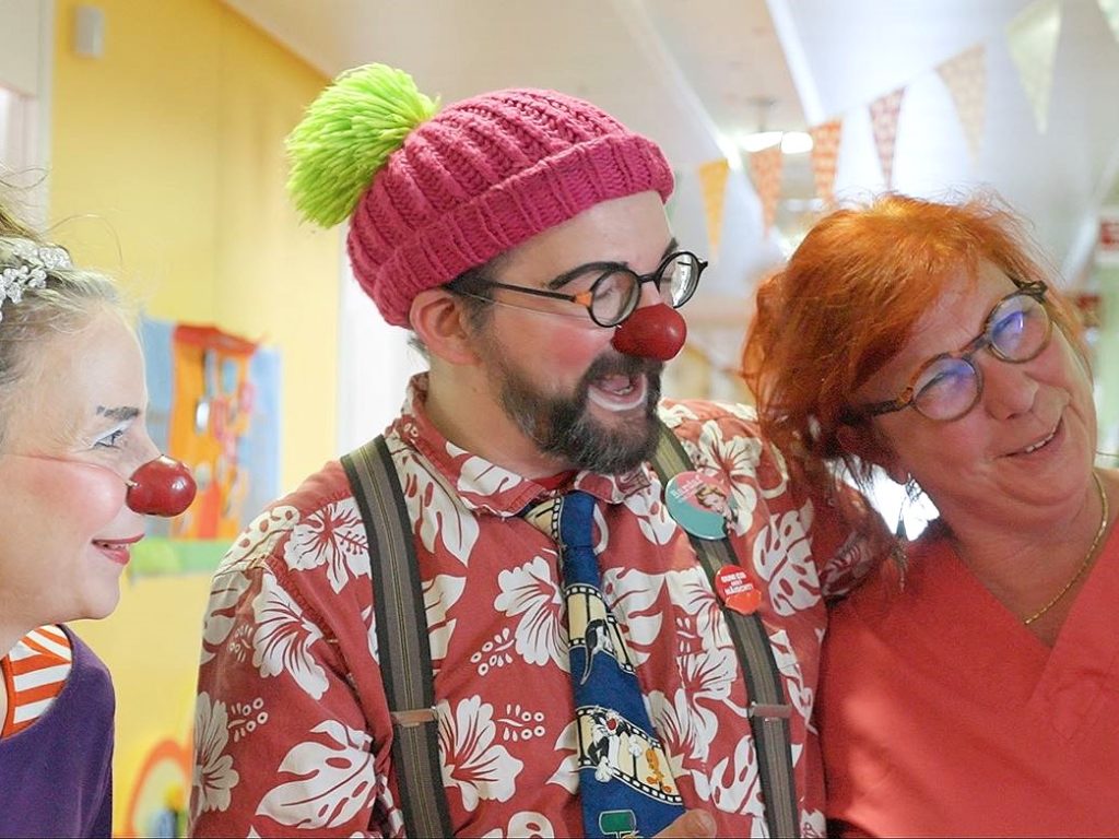 Trois adultes sourient et rient ensemble. Deux d'entre eux sont habillés en clowns avec un nez rouge, l'autre porte un bonnet en tricot rose et une chemise à fleurs, tandis que la troisième personne, en blouse rouge, se tient à côté d'eux. L'arrière-plan est lumineux et joyeux.