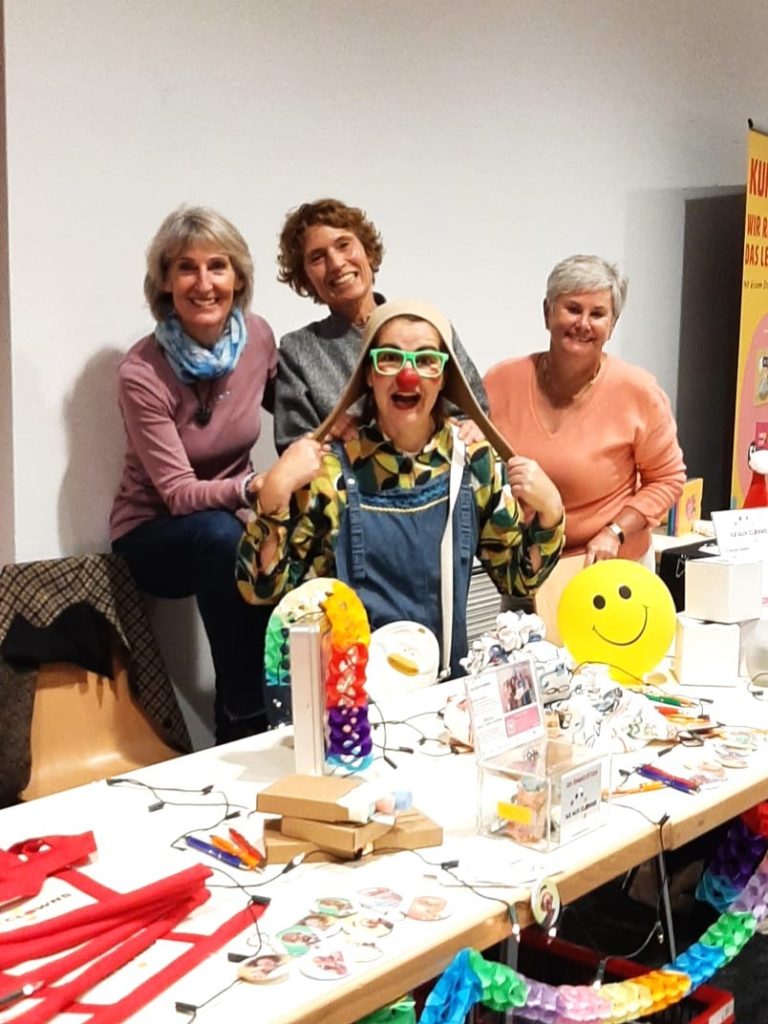 Quatre femmes sourient à une table couverte d'objets artisanaux, de jouets et de décorations colorés. L'une d'entre elles porte des lunettes amusantes et tient un foulard au-dessus de sa tête. La scène est joyeuse et créative.