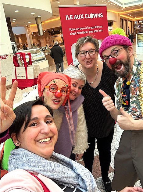 Cinq personnes, dont deux déguisées en clowns avec des nez rouges, des chapeaux colorés et des vêtements brillants, sourient et posent pour un selfie dans un centre commercial. Un panneau rouge ILE AUX CLOWNS est visible à l'arrière-plan.