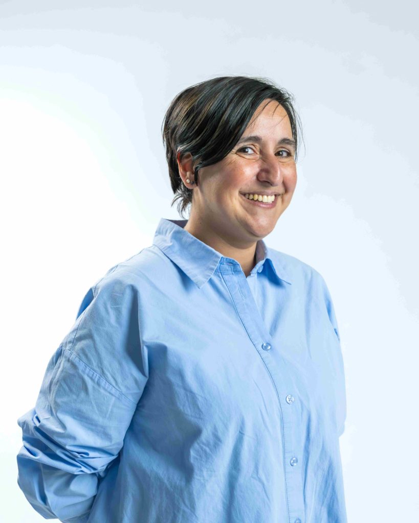 Une personne aux cheveux courts et foncés, vêtue d'une chemise à boutons bleu clair, se tient debout, souriante, les mains derrière le dos, sur un fond uni et clair.