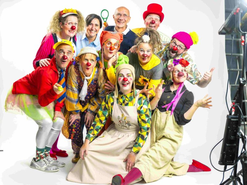 Un groupe joyeux d'adultes portant des costumes de clowns colorés et des nez rouges pose ensemble, souriant et faisant des gestes ludiques devant un arrière-plan blanc. Les lumières et l'équipement du studio sont visibles sur la droite.