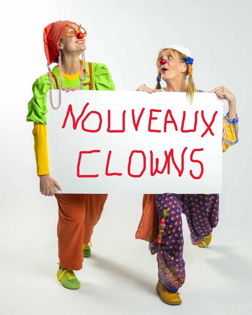 Deux clowns colorés sourient et regardent en l'air tout en tenant un grand panneau blanc avec un texte rouge peint à la main indiquant NOUVEAUX CLOWNS sur un fond blanc uni.