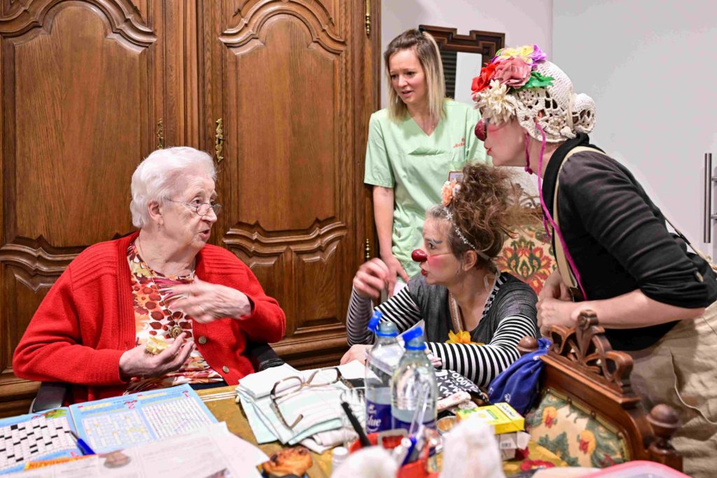 Une femme âgée vêtue d'un pull-over rouge discute avec deux personnes déguisées en clowns et une aide-soignante en uniforme vert autour d'une table couverte de papiers et de bouteilles dans une pièce confortable.