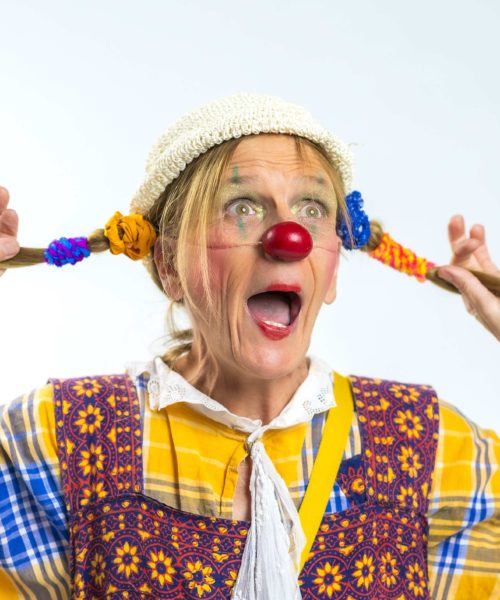 Une femme déguisée en clown, avec un nez rouge, un visage peint en couleurs et des tresses, porte une robe à motifs et un bonnet en tricot. Elle tire ses tresses sur les côtés et affiche une expression de surprise.