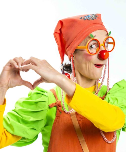 Une personne habillée en clown, avec des vêtements vert et orange, des lunettes orange et un nez rouge, dessine un cœur avec ses mains en souriant.