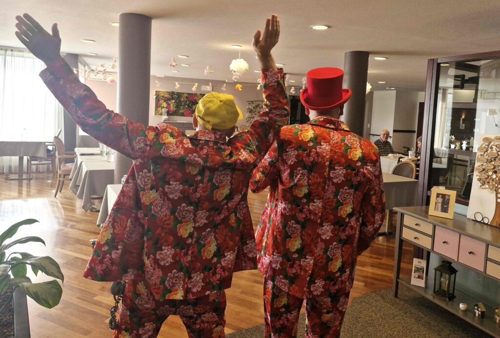 Deux personnes vêtues de costumes à fleurs assortis, l'une avec un chapeau jaune et l'autre avec un haut-de-forme rouge, se tiennent dos à la caméra dans une pièce très éclairée avec des tables et des planchers en bois.