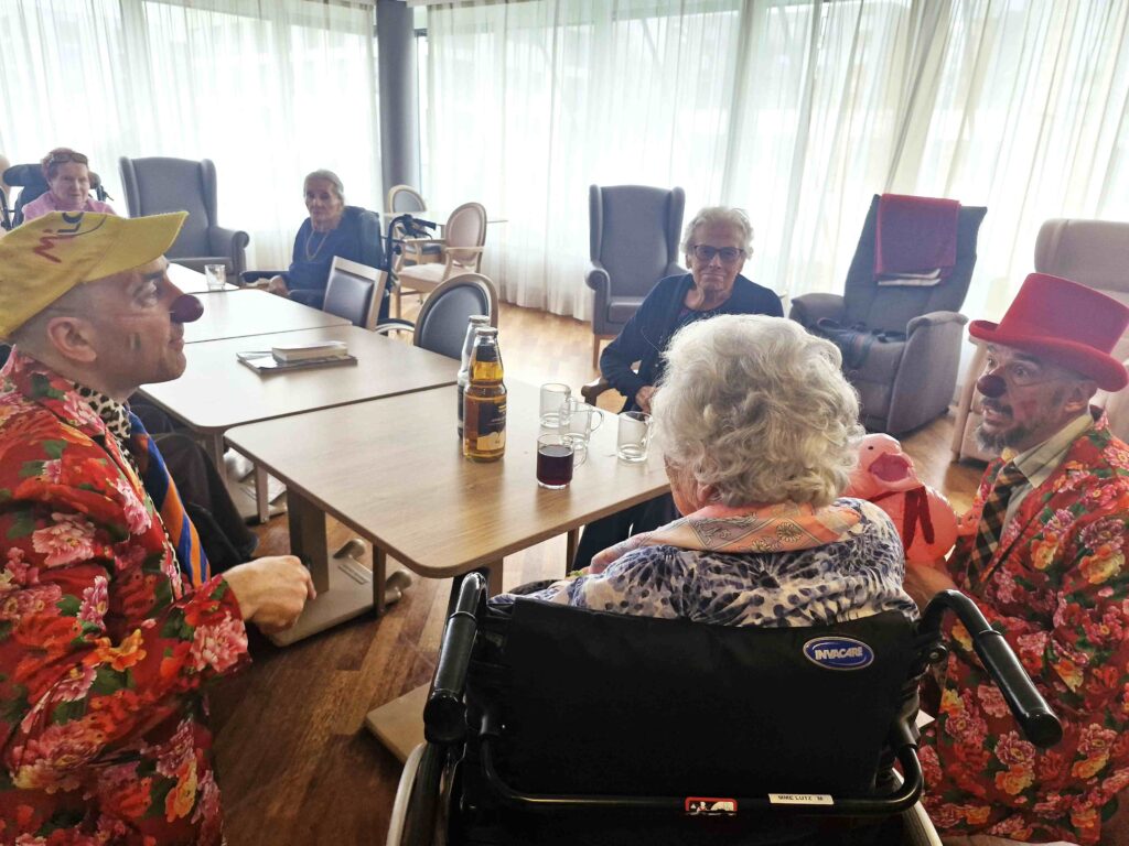 Deux clowns en costumes colorés divertissent des personnes âgées assises autour d'une table dans une pièce lumineuse. Une femme âgée est en fauteuil roulant et il y a des boissons et des livres sur la table.