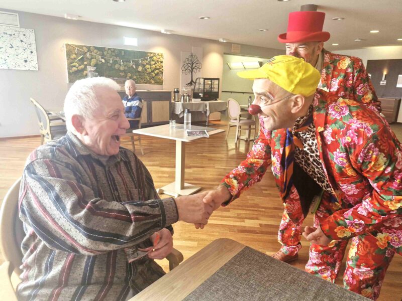 Un homme âgé assis à une table serre la main d'un clown souriant aux vêtements colorés et au bonnet jaune, tandis qu'un autre clown au bonnet rouge se tient derrière eux. Ils se trouvent dans une pièce claire et spacieuse.