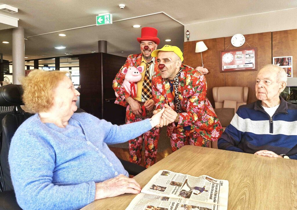 Deux personnes âgées sont assises à une table avec un journal, tandis que deux clowns en tenues colorées et au nez rouge interagissent avec elles, tenant un cochon en peluche et créant une atmosphère joyeuse dans une pièce lumineuse.