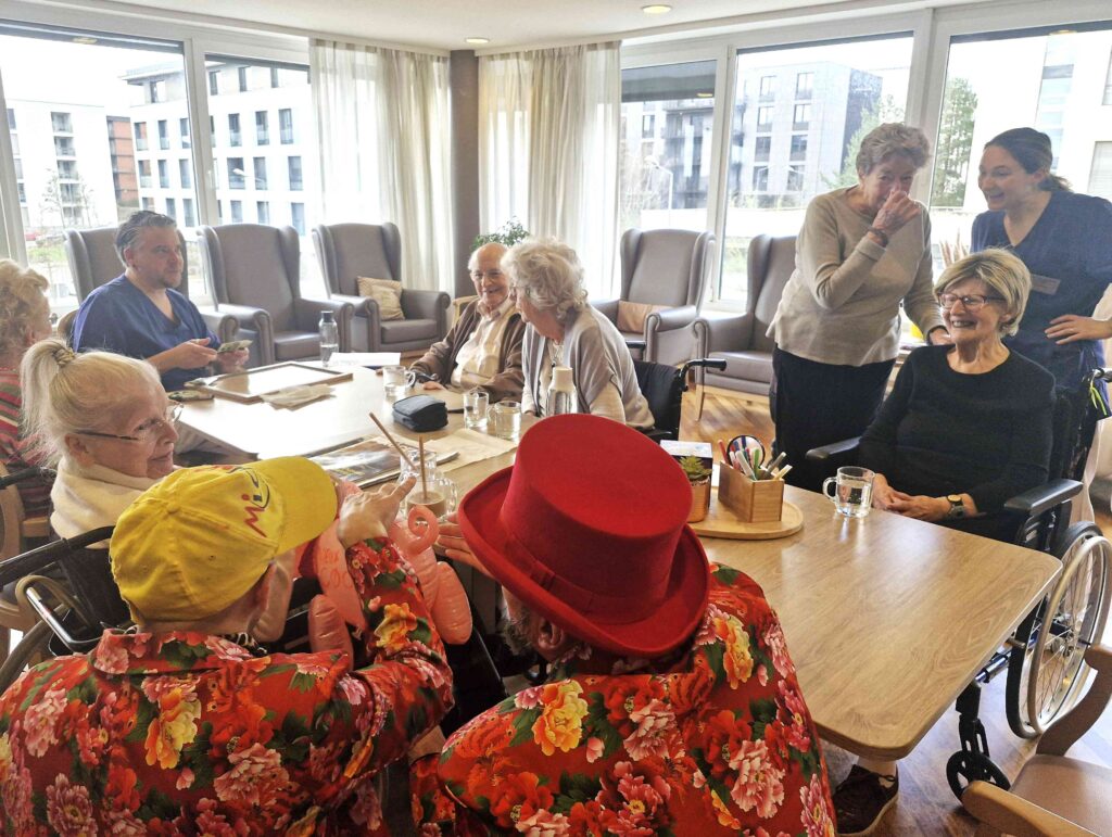 Un groupe de personnes âgées est assis autour de tables dans une pièce lumineuse, souriant et discutant. Deux personnes portant des vêtements et des chapeaux colorés, peut-être des artistes, sont assises au premier plan. L'atmosphère est animée et joyeuse.