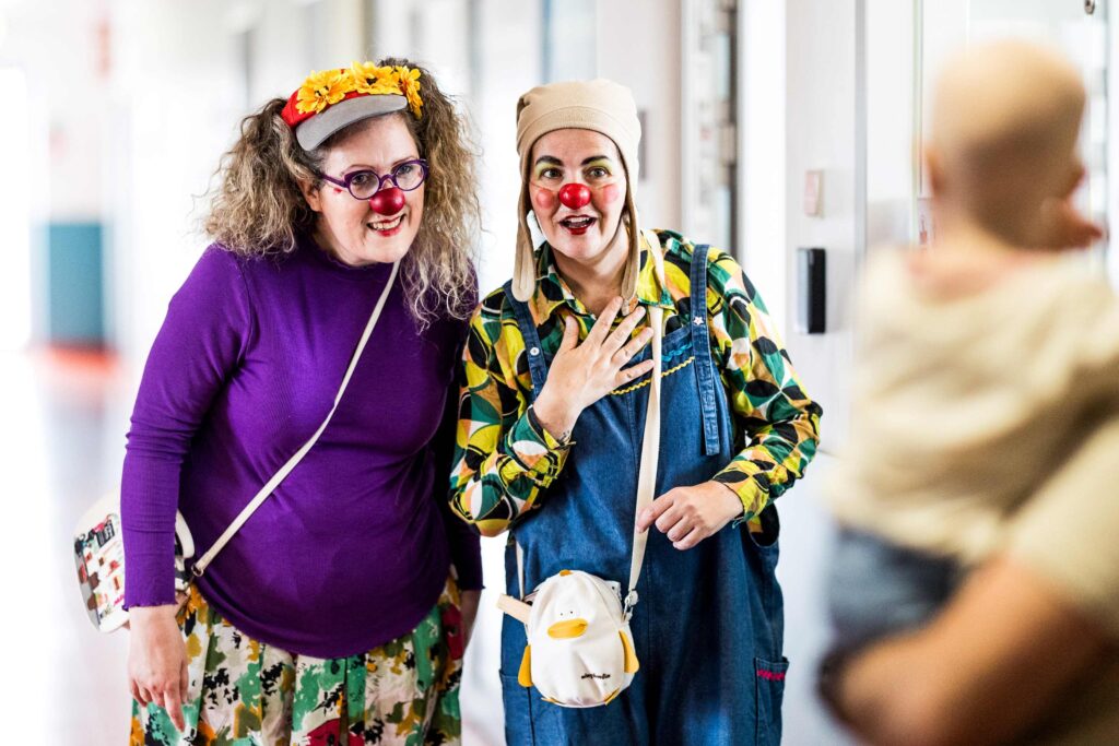 Deux clowns au nez rouge et aux tenues colorées sourient et interagissent avec une silhouette floue tenant un bébé dans un couloir lumineux. L'un des clowns porte une couronne de fleurs, l'autre un chapeau beige et une salopette en jean.