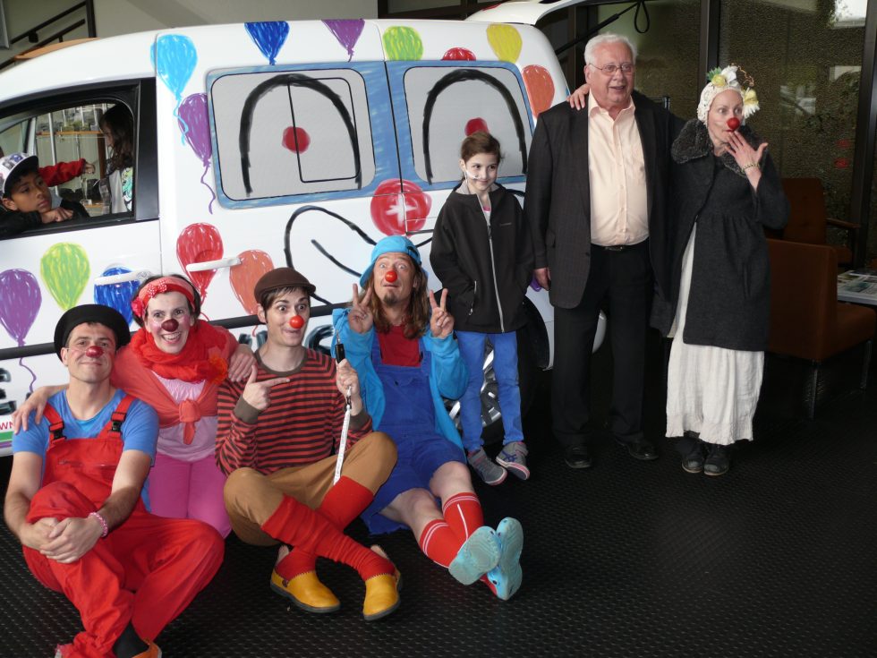 Un groupe de clowns en costumes colorés pose et sourit avec trois adultes et un enfant devant une camionnette décorée d'un visage de clown et de ballons.