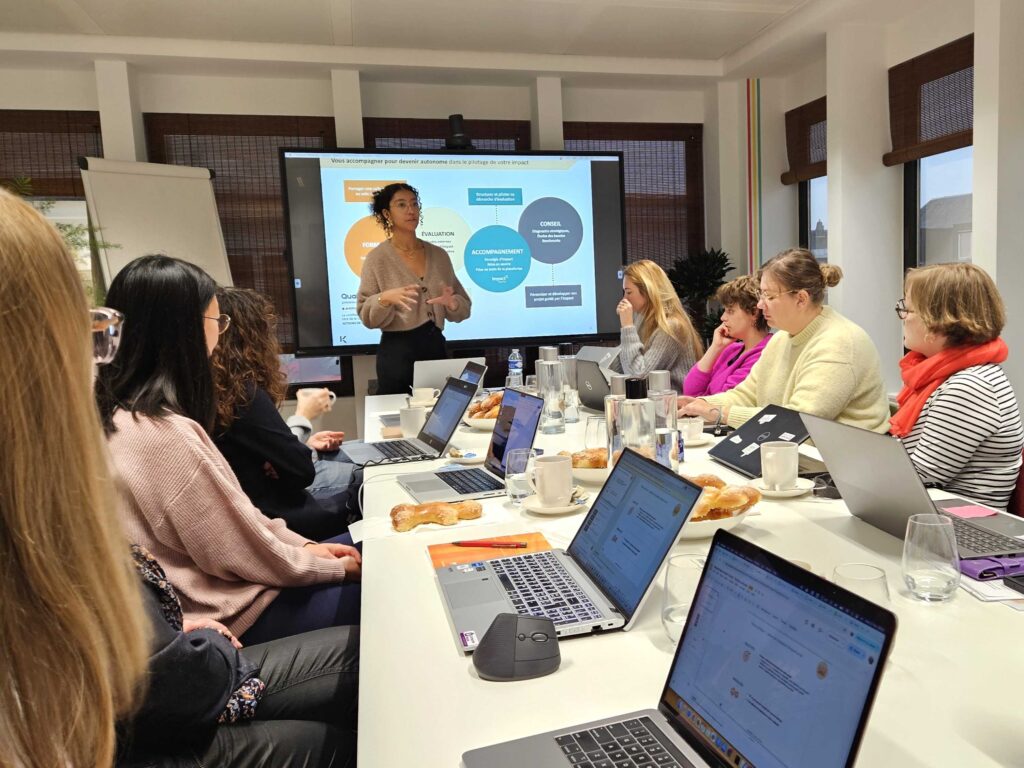 Une femme se présente devant un écran affichant un organigramme à un groupe de personnes assises autour d'une table de conférence avec des ordinateurs portables, des cahiers, des tasses de café et des pâtisseries.